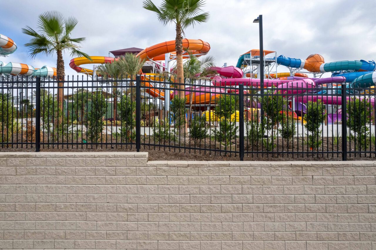Water park exterior view with colorful spiraling slides in orange, pink, purple, and teal, seen beyond a black metal fence atop a tan concrete block retaining wall, with palm trees and young shrubs planted along the perimeter.