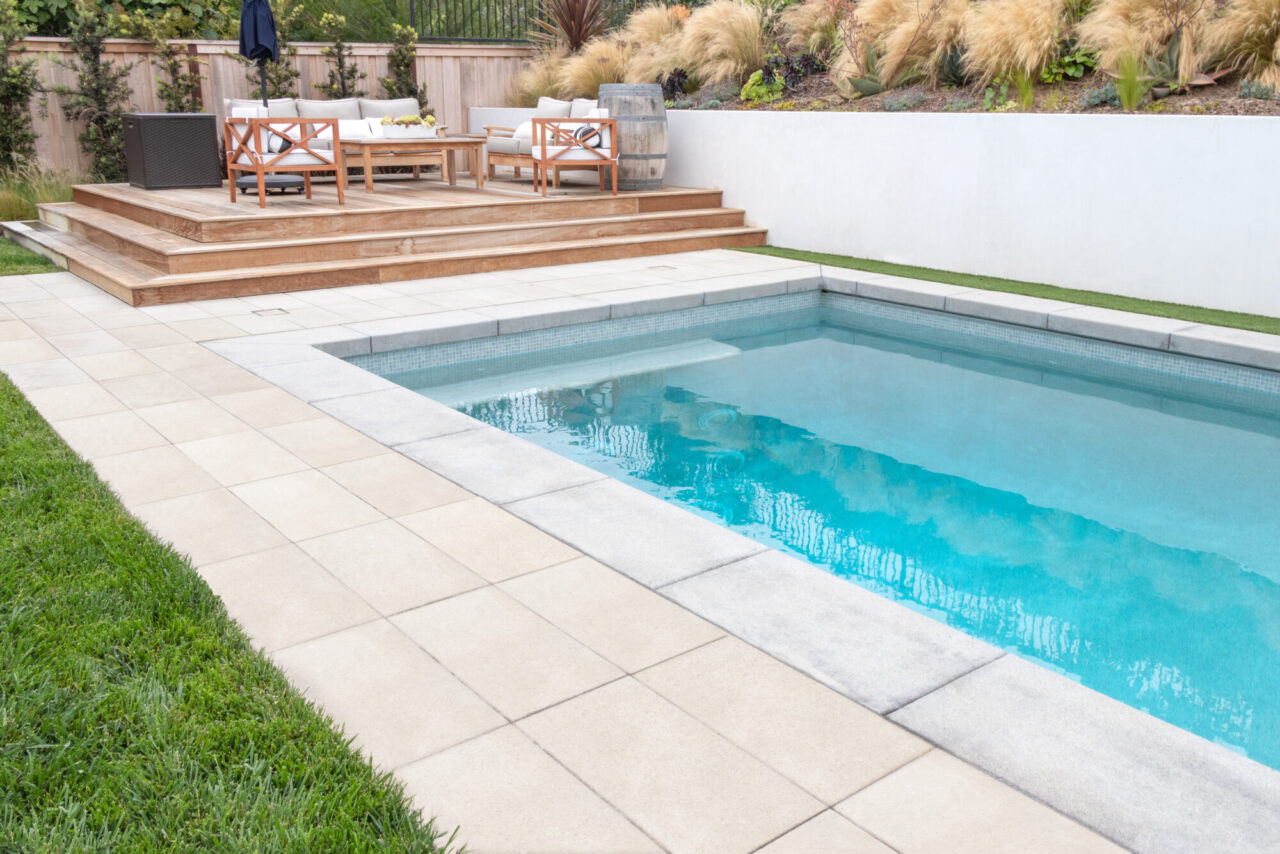 Rectangular pool with a paver deck bordered by a raised wood deck with outdoor seating and a fenced garden.