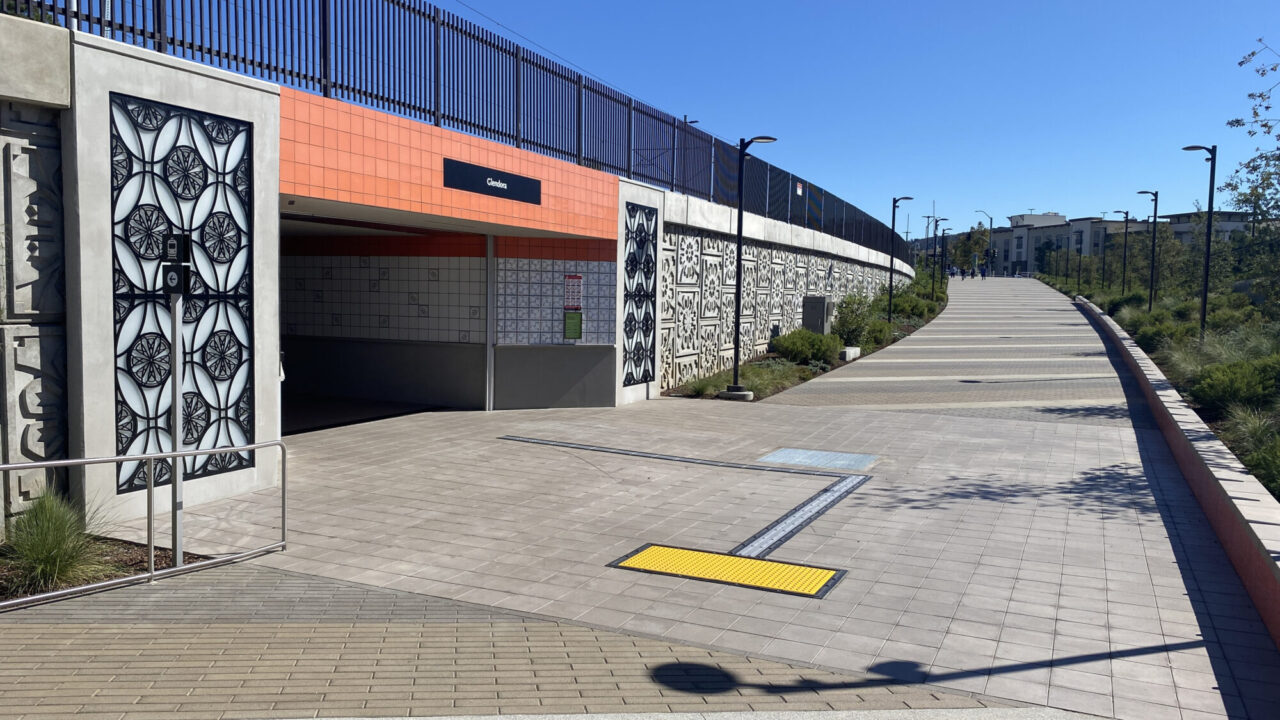 Decorative concrete wall panel featuring repeating carved medallion patterns, paired with a matching black metal circular screen gate, bordered by ornamental grasses and paver walkways in a modern outdoor setting.