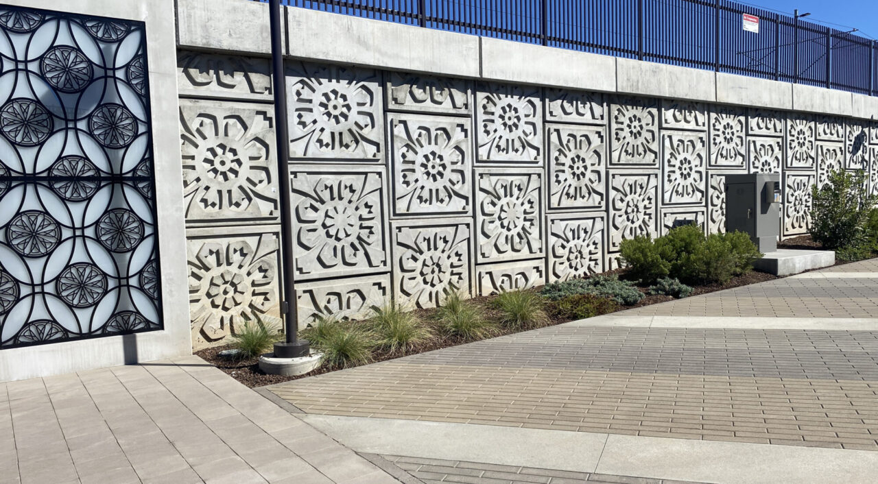 Decorative concrete wall panel featuring repeating carved medallion patterns, paired with a matching black metal circular screen gate, bordered by ornamental grasses and paver walkways in a modern outdoor setting.