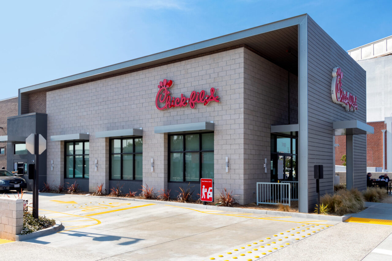 Modern Chick-fil-A restaurant exterior with light gray concrete block and metal wall paneling, angled roof overhang, storefront windows, drought-tolerant landscaping, and a drive-thru lane with painted directional markings.