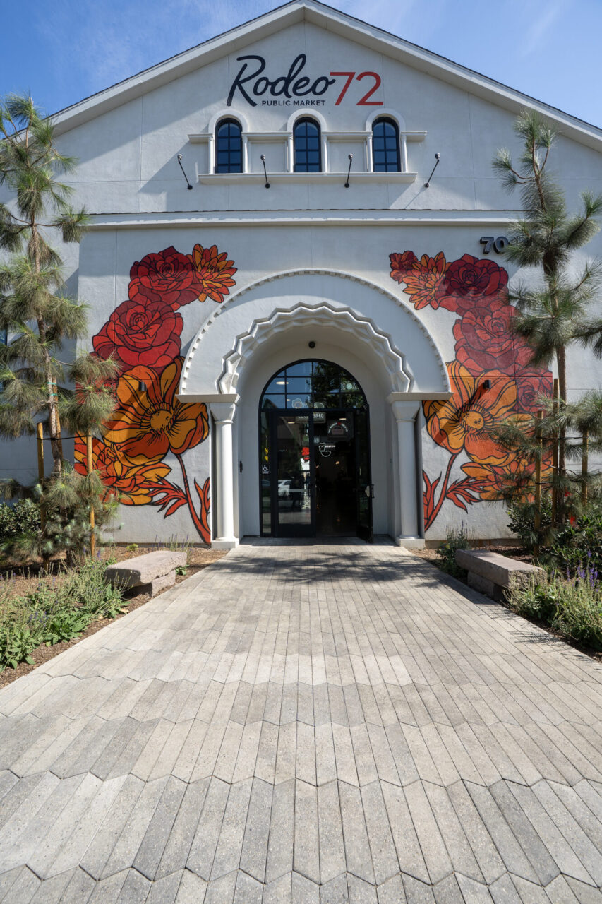Rodeo 72 Public Market entrance with a white stucco arched doorway framed by colorful red and orange floral mural art, leading up a chevron-patterned paver walkway lined with young pine trees and landscaping.