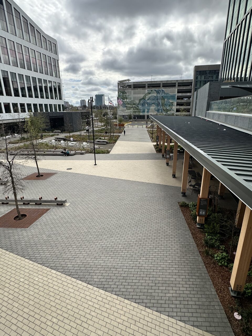 Modern urban plaza with gray paver flooring, wood-and-steel bench seating arranged around planted trees, an elevated glass skywalk bridge, and multi-story glass office buildings in the background.