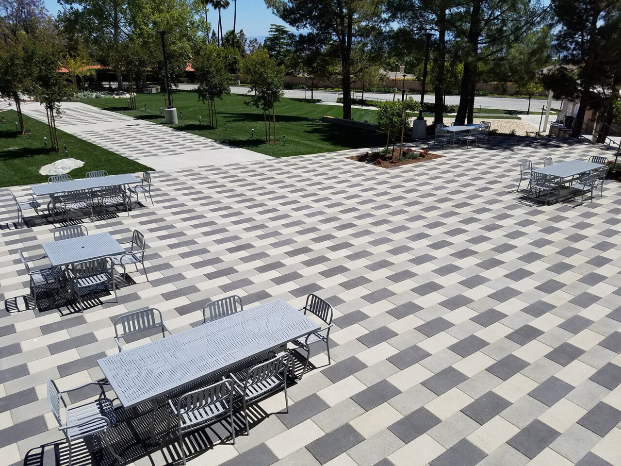 Aerial view of a church campus courtyard with a checkerboard-patterned paver plaza, gray metal picnic tables and chairs, bordered by tree-lined pathways and open lawn areas.