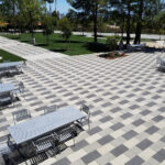 Aerial view of a church campus courtyard with a checkerboard-patterned paver plaza, gray metal picnic tables and chairs, bordered by tree-lined pathways and open lawn areas.