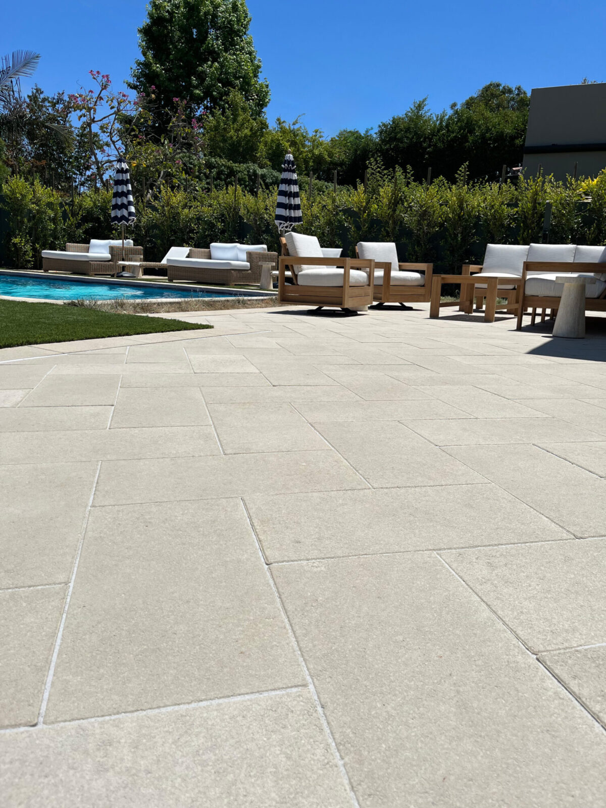 Light paver patio with wooden lounge seating and striped umbrellas beside a pool bordered by hedges.