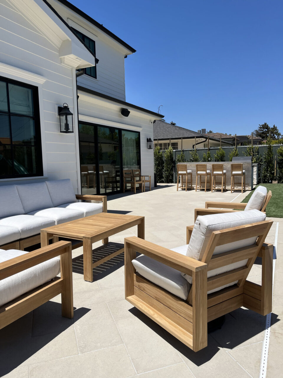 Backyard patio with teak furniture and cushioned seating set beside a white farmhouse-style home with an outdoor bar and hedges in the background.