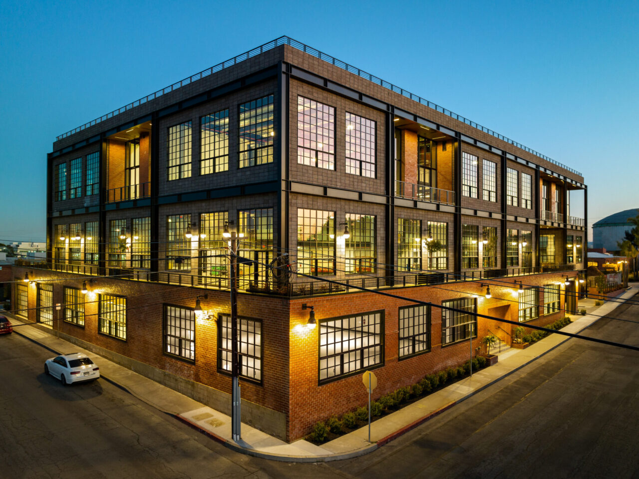 Three-story industrial-style loft building at dusk with red brick lower level and gray concrete masonry upper floors, large black-framed factory windows glowing warmly from within, and exterior sconce lighting along a corner street setting.