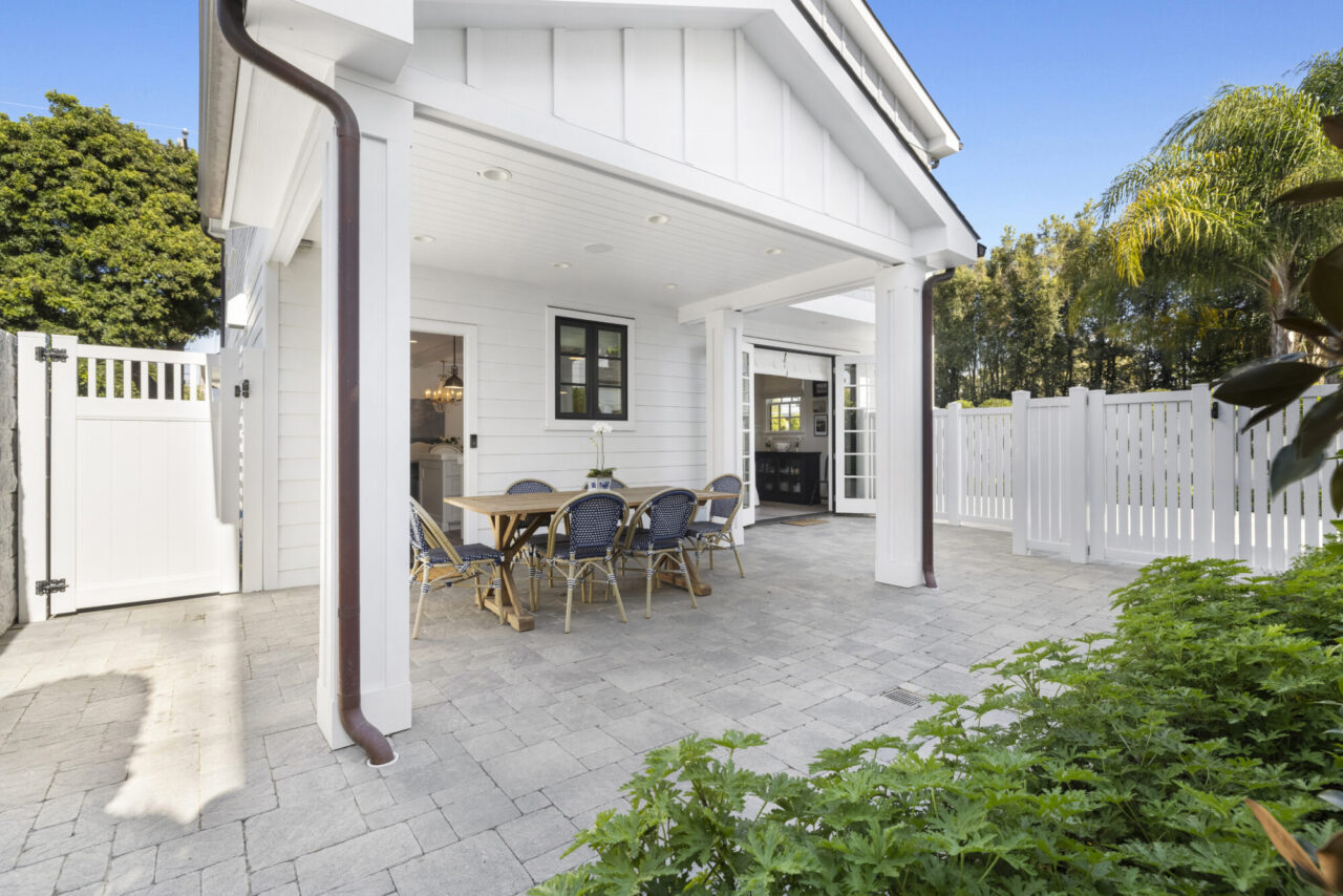 Covered paver patio with a wooden dining table and blue chairs, set beside a white farmhouse-style home with a fenced yard.