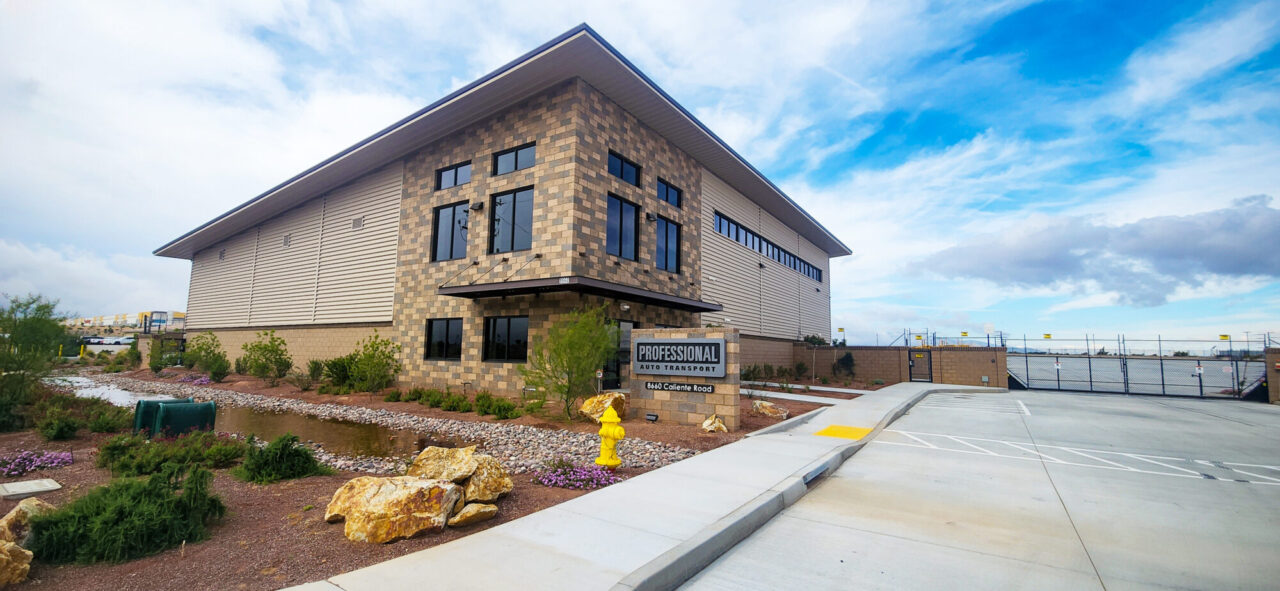 Modern two-story commercial building with beige and brown concrete block walls, metal siding, and large windows, featuring a "Professional Auto Transport" sign at 8660 Caliente Road, with desert landscaping, boulders, and a paved parking lot in the foreground under a partly cloudy sky.