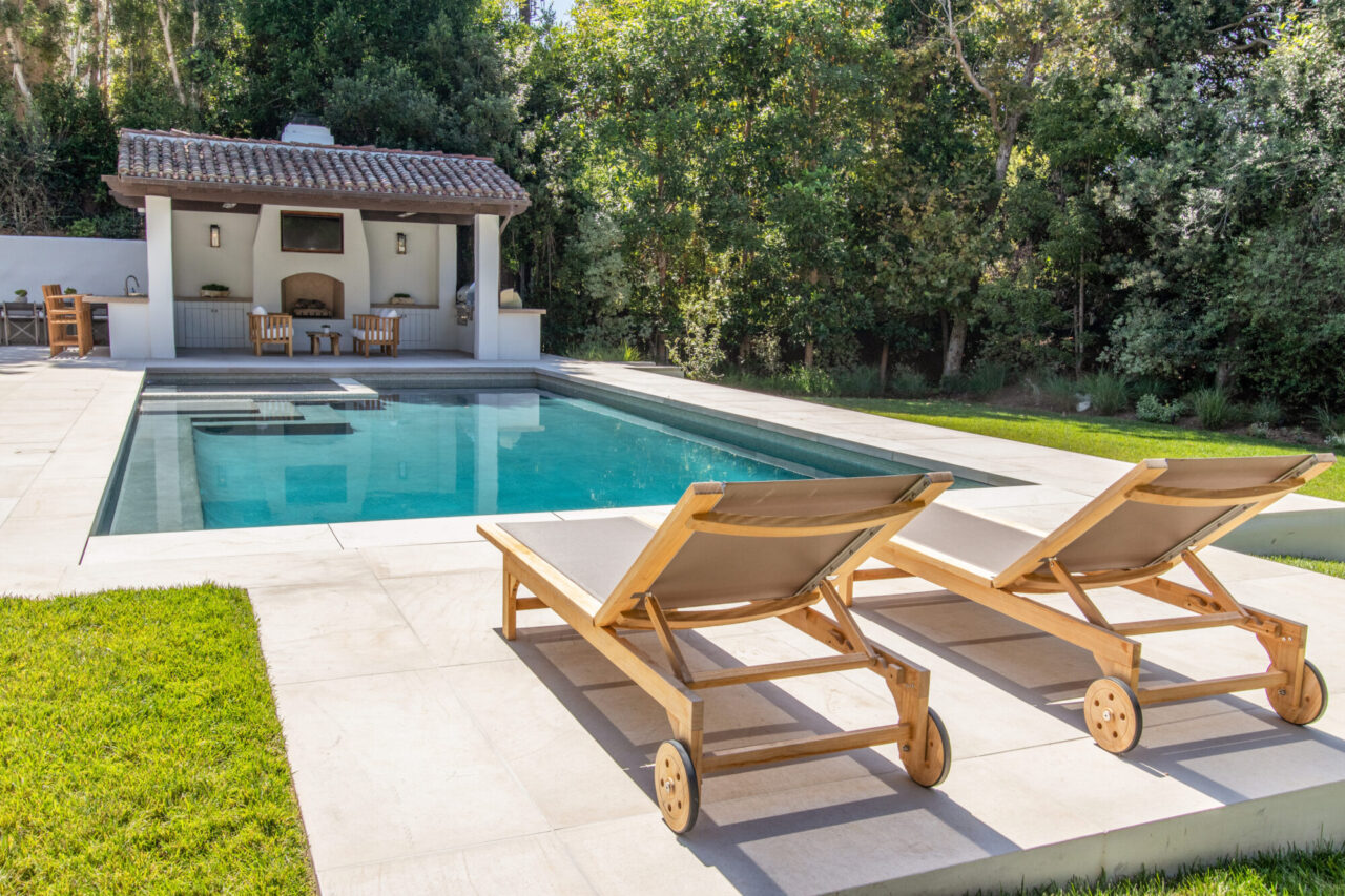 Wooden lounge chairs on a natural stone pool deck with a tiled-roof pool house and fireplace.