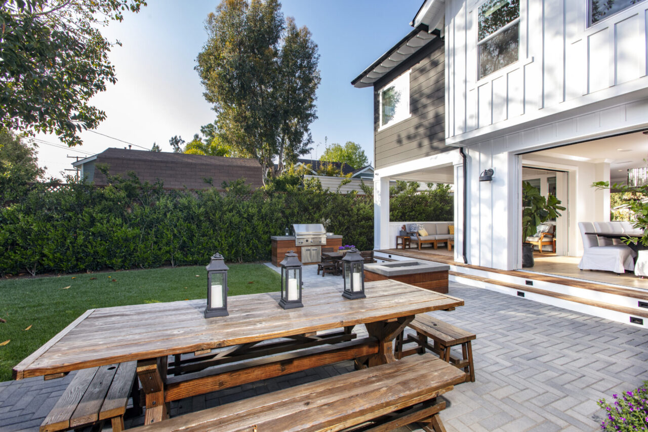 Backyard paver patio with a wooden picnic table and lanterns, featuring an outdoor kitchen and open sliding doors into a white farmhouse-style home.