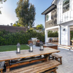 Backyard paver patio with a wooden picnic table and lanterns, featuring an outdoor kitchen and open sliding doors into a white farmhouse-style home.