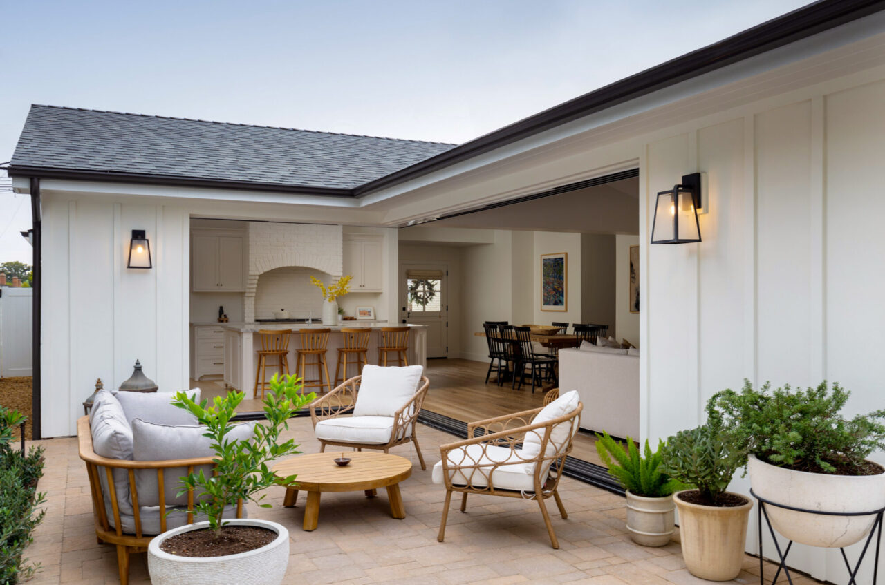 Outdoor paver patio with rattan seating and potted plants, opening through folding glass doors into a white farmhouse-style kitchen and dining area.