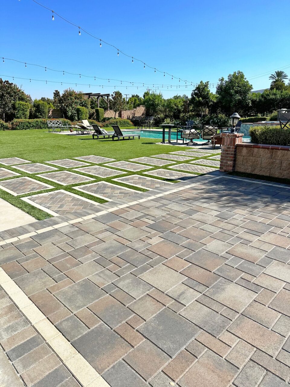Backyard paver patio with a diamond-pattern grass inlay, leading to a pool area with lounge chairs and string lights overhead.