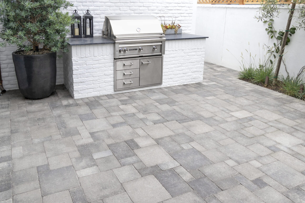 Built-in stainless steel grill station with painted brick base on a gray paver patio, flanked by potted greenery.
