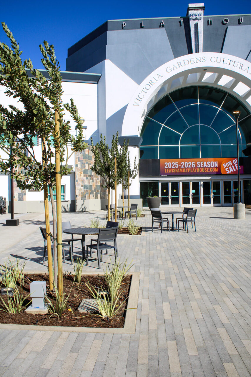 Victoria Gardens Cultural Center plaza with an arched glass entrance to the Lewis Family Playhouse, outdoor cafe tables and chairs, young staked trees in planting beds, and gray paver flooring under a clear blue sky.