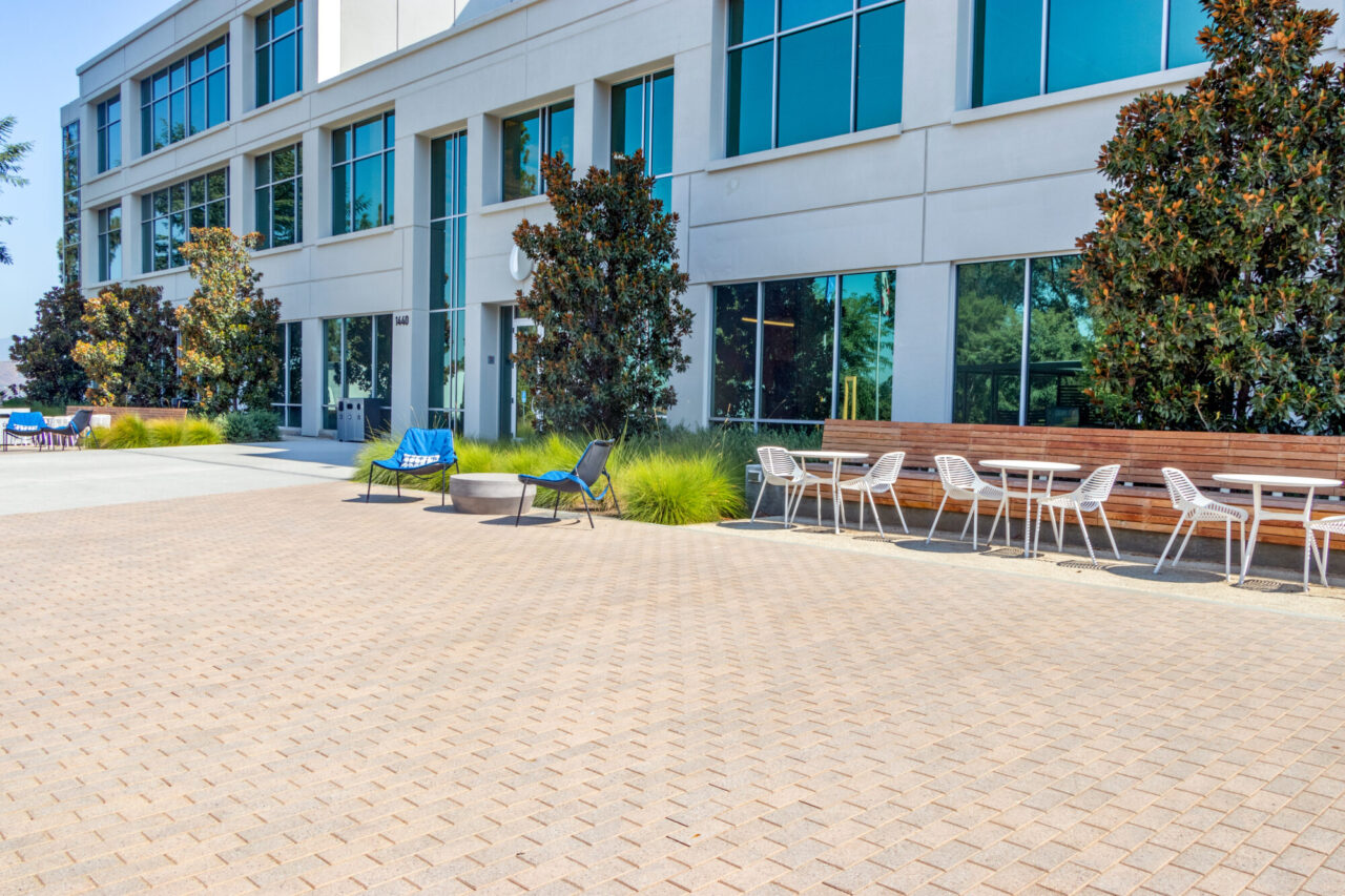 Corporate office building exterior at 1440 with a glass curtain-wall facade, outdoor seating area featuring blue lounge chairs, white bistro tables, a wood accent bench, ornamental grasses and paver plaza.