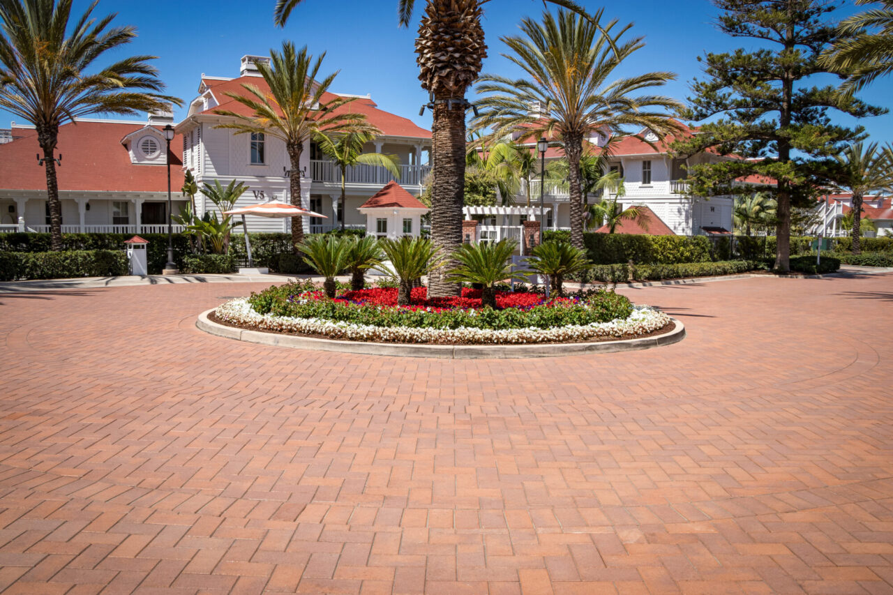 Circular paver courtyard with a landscaped center island of palm trees and vibrant red and white flowers, fronted by red-roofed white Victorian resort buildings and shade trees.