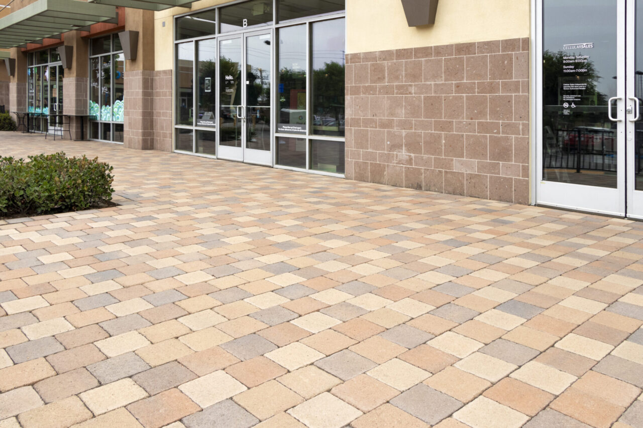 Retail shopping center storefronts with multicolored tan and brown paver walkway, glass entrance doors, and tile-clad exterior walls, including a visible cell phone storefront sign.