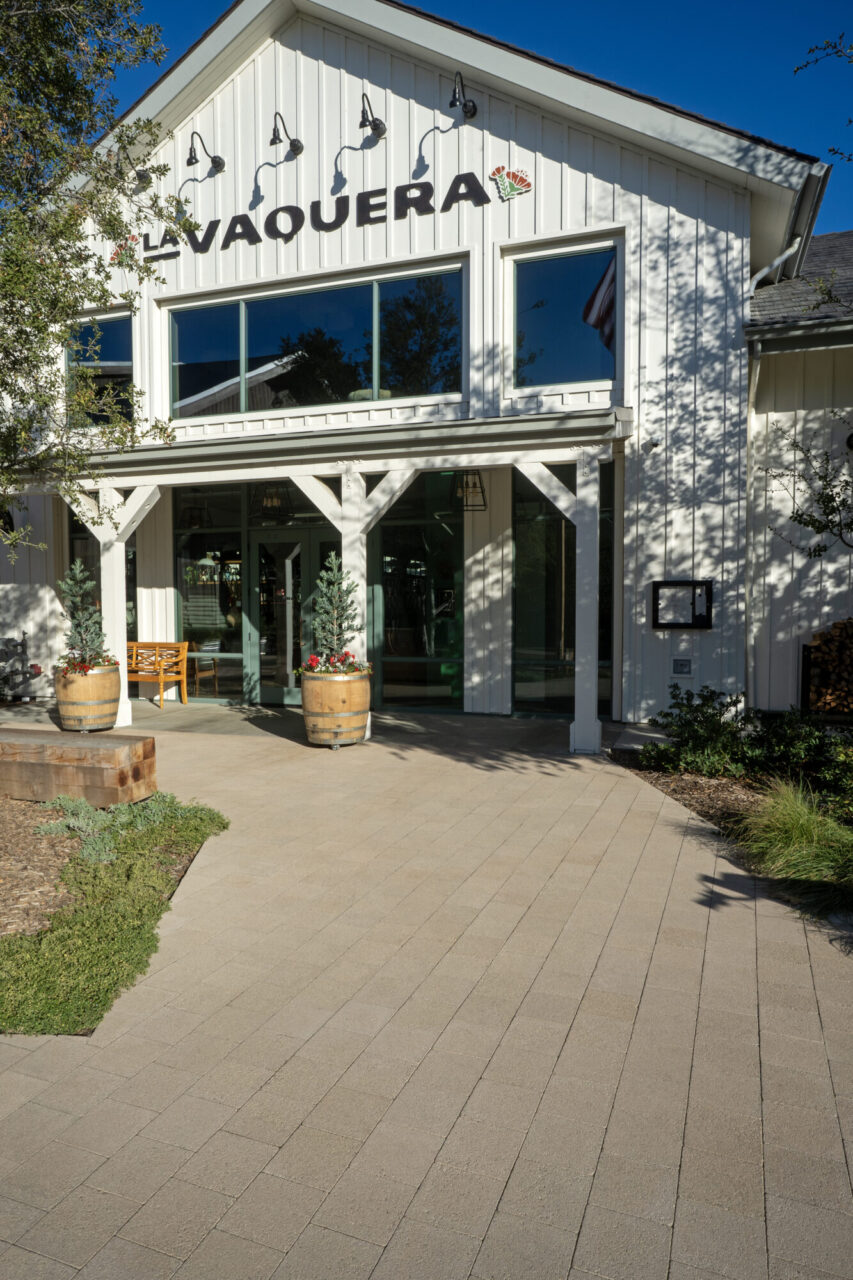 "La Vaquera" restaurant entrance with white board-and-batten siding, wood post porch, wine barrel planters with topiary trees, sconce lighting, and a paver walkway leading to the glass storefront entrance.
