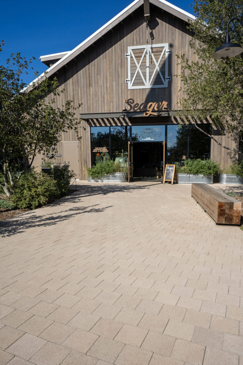 Barn-style storefront with vertical wood siding, a decorative white shutter accent, and "Seager" signage above the entrance, fronted by a wood beam bench, metal planters, and a paver plaza.