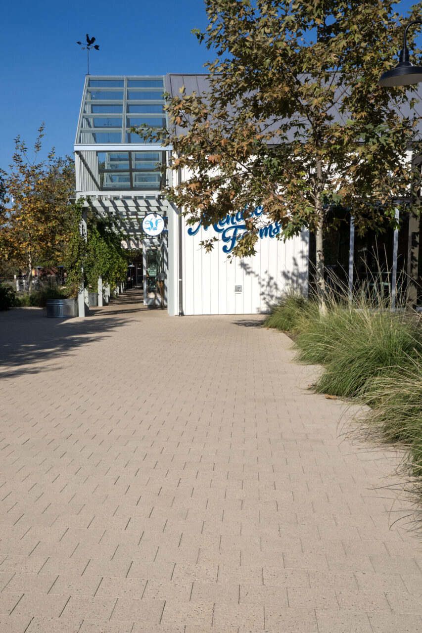 Storefront building with a cursive "Mendocino Farms" sign and glass tower entrance topped by a weathervane, set along a paver walkway lined with ornamental grasses and shade trees.