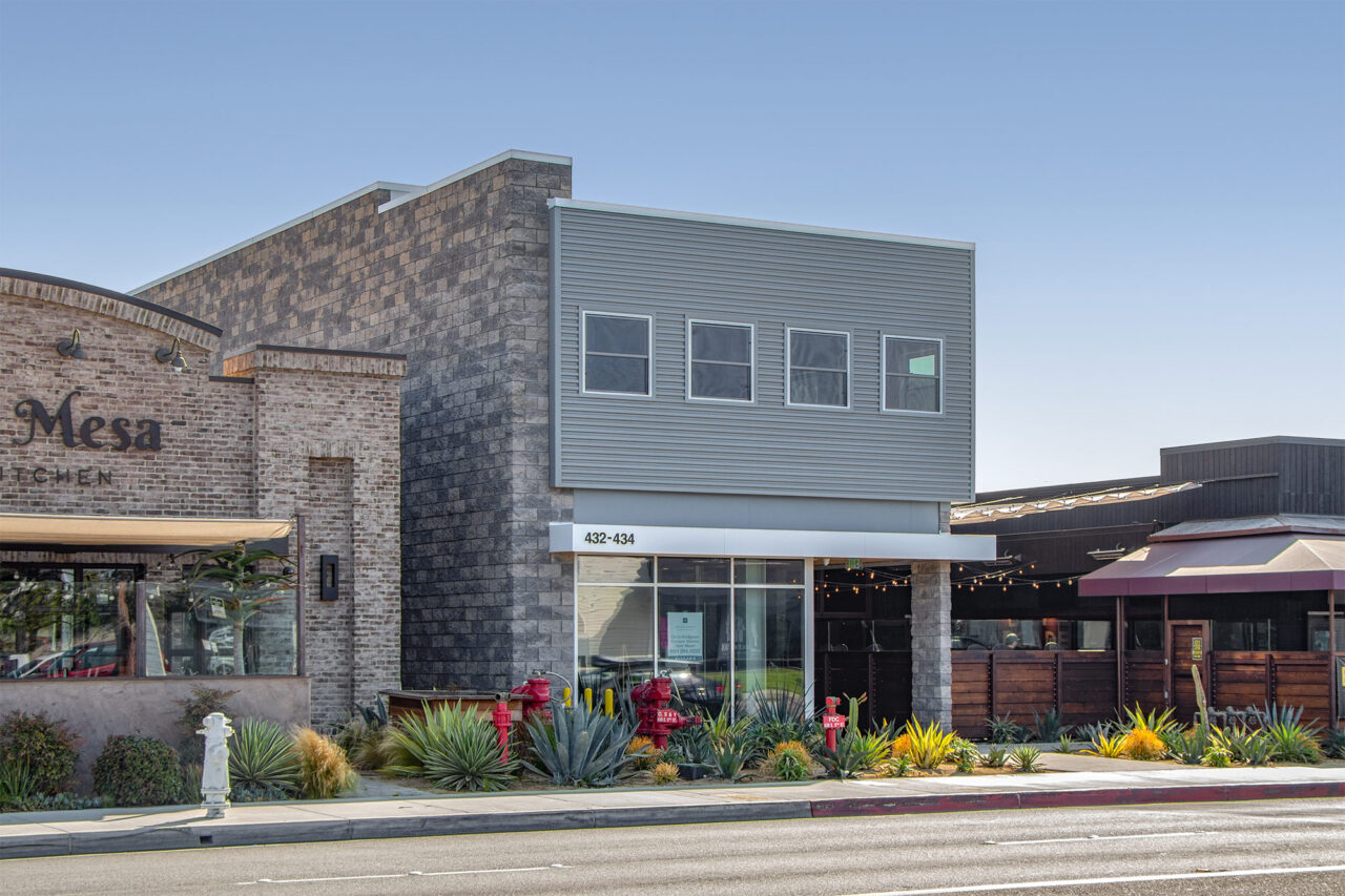 Two-story commercial storefront building at 432-434 with gray metal siding above and brick and block masonry below, storefront glass windows, and a street-facing garden bed of agave, succulents, and ornamental grasses.