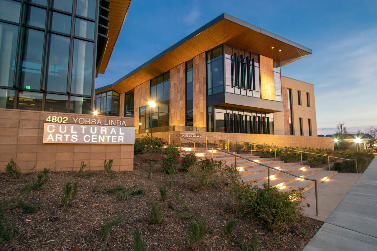 Yorba Linda Cultural Arts Center building at dusk with warm wood-clad overhangs, glass curtain walls, and stone facade, featuring an illuminated staircase with metal handrails leading up through a landscaped, drought-tolerant garden bed.