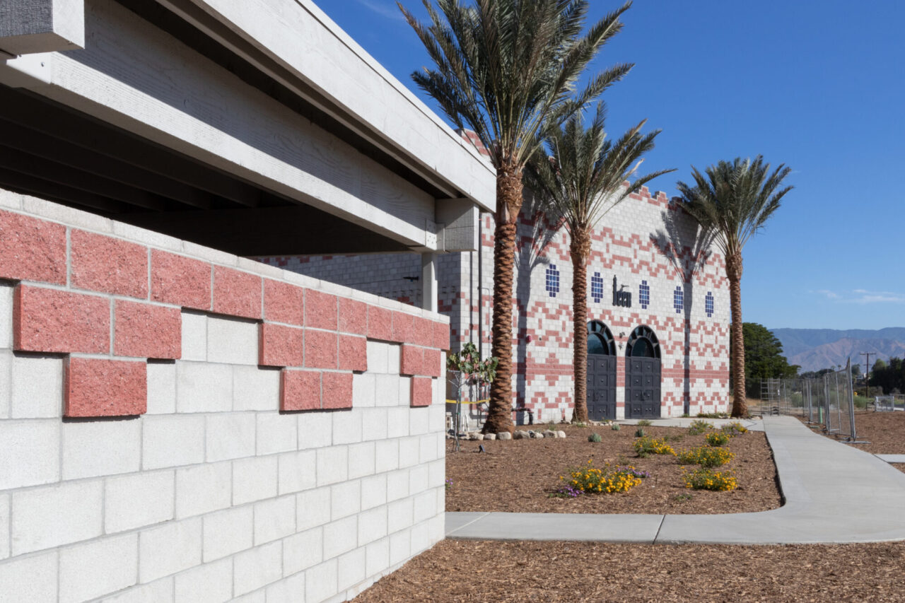 Contemporary desert building with red and white patterned block walls, arched double doors, and tall palm trees, set against a xeriscaped garden with wildflowers and distant mountains.