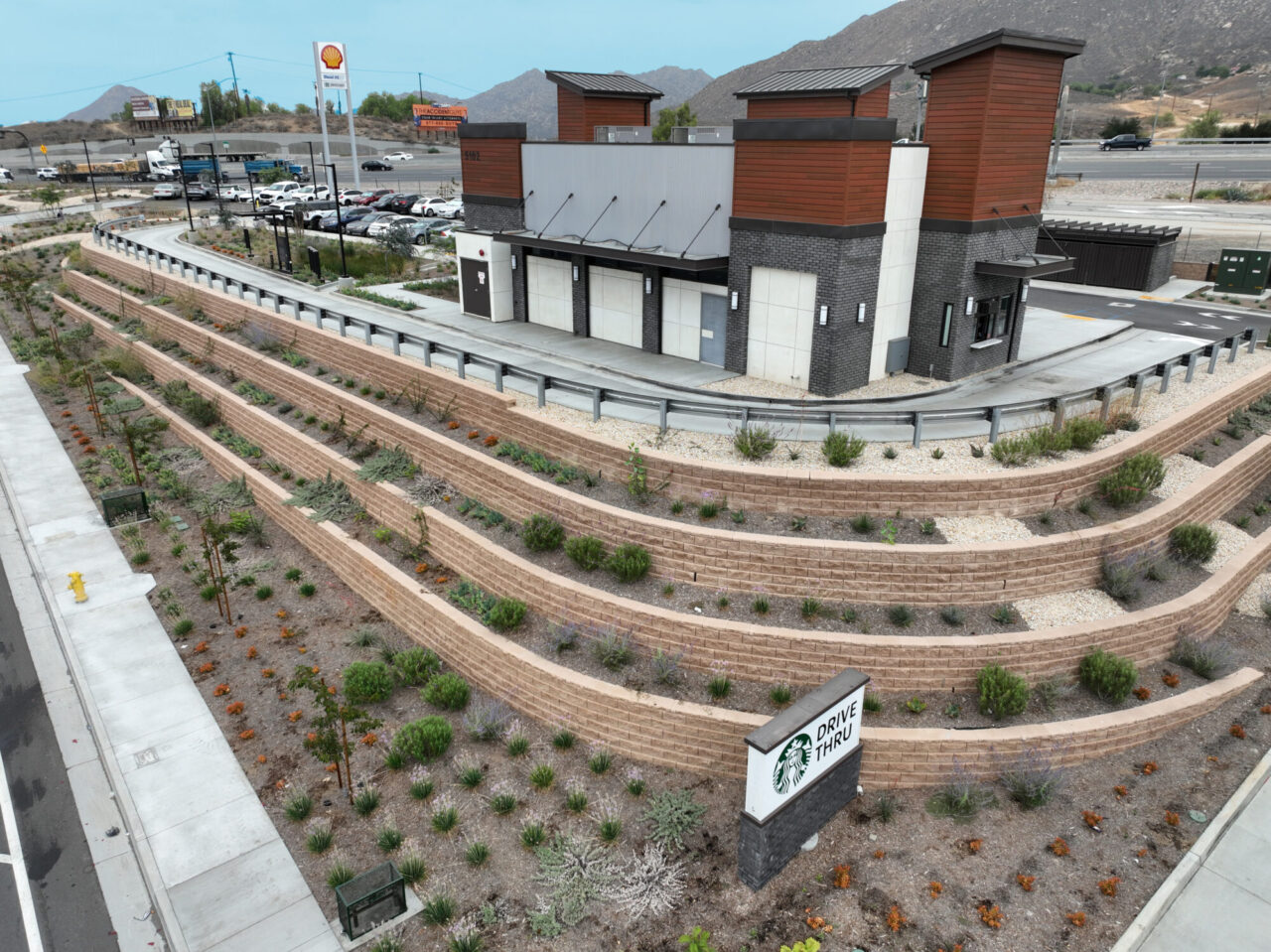 Aerial view of a Starbucks drive-thru building with dark tile and wood-paneled towers, situated above a terraced retaining wall landscape with desert plants, a "Drive Thru" sign in the foreground, and a parking lot, road, and distant mountains visible in the background.