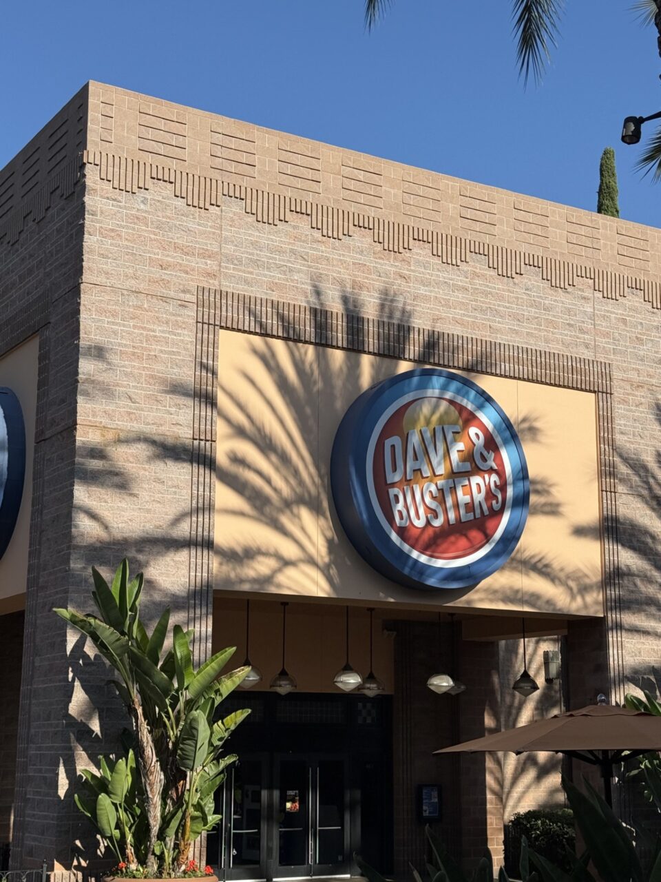 Exterior view of a Dave & Buster's storefront with a circular blue and red logo sign above the entrance, tan concrete block structures with decorative trim, glass double doors, hanging pendant lights, and palm tree shadows cast across the building.
