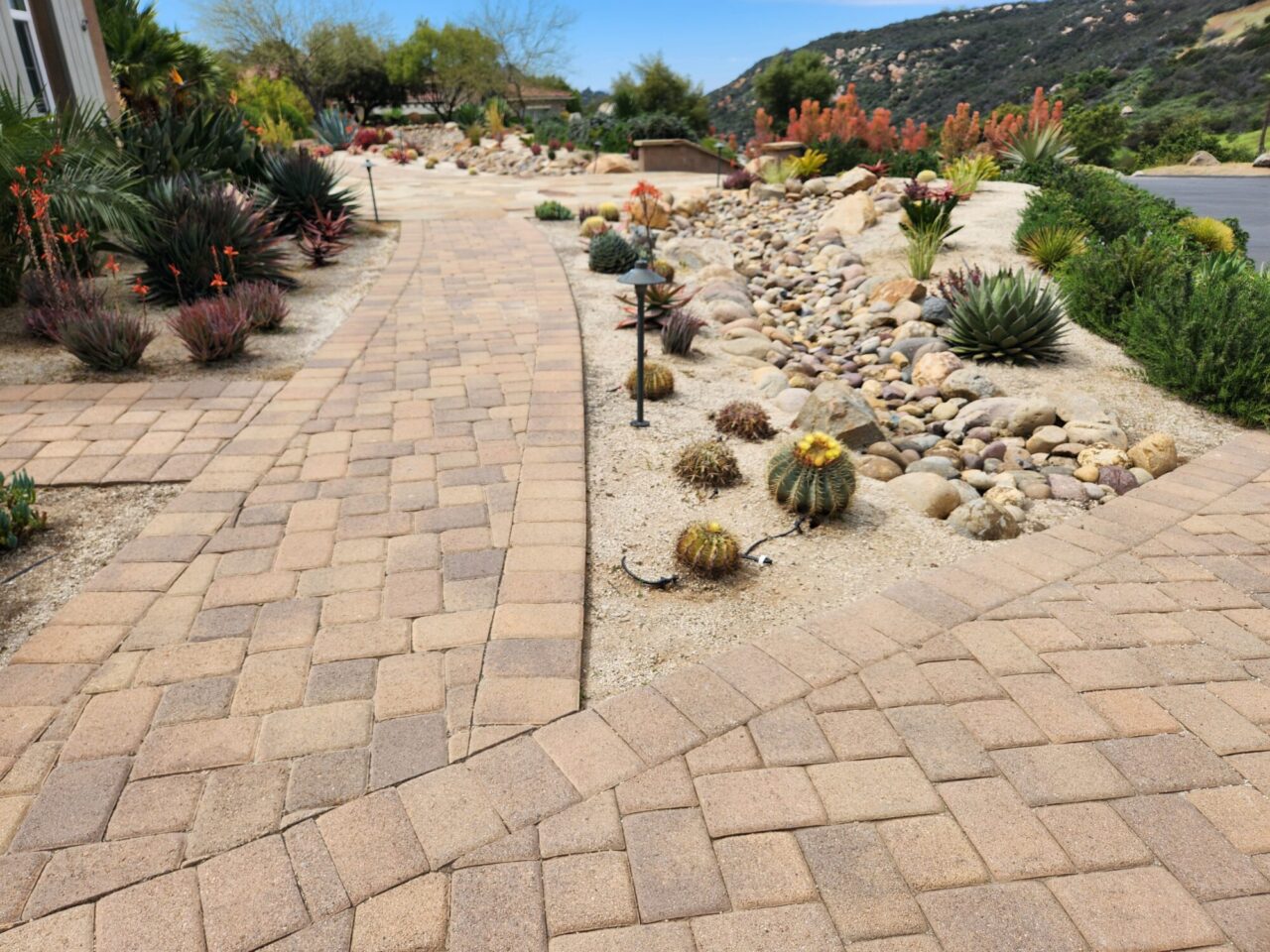 Paver walkway winding past a dry riverbed rock garden with barrel cactus and desert plants framed by hills in the background.
