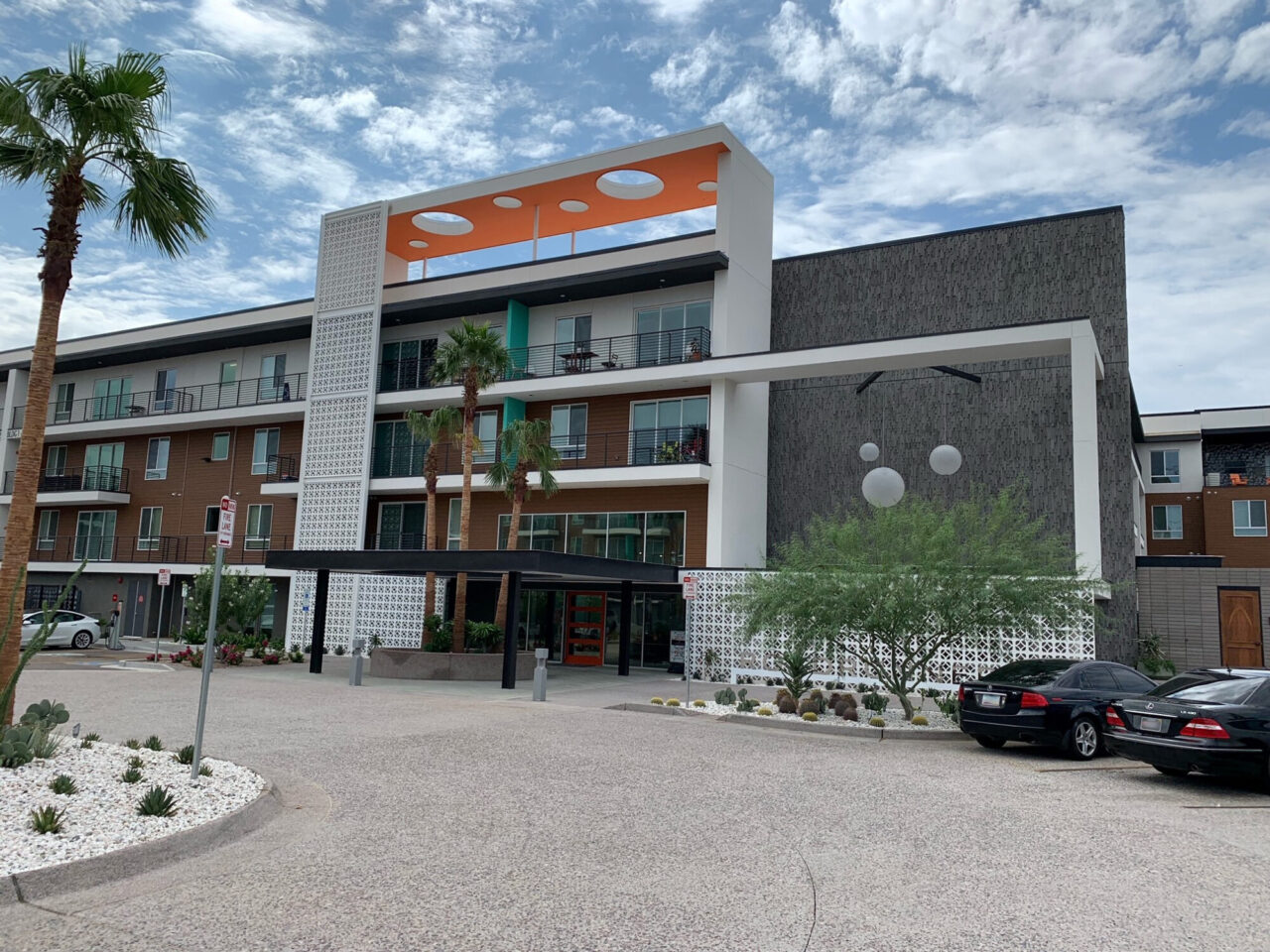 Multi-story mid-century modern style apartment complex with brown and white paneling, a rooftop orange accent with circular cutouts, decorative white breeze block walls, hanging globe lights, and a red-doored entrance, with palm trees, desert landscaping, and a parking area in the foreground.
