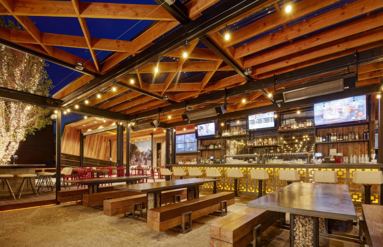 Nighttime view of an outdoor bar and dining area with an exposed timber pergola roof strung with pendant lights, a fully stocked bar backlit with amber decorative panels, mounted TVs, wooden communal tables with benches, and string-lit trees in the background.