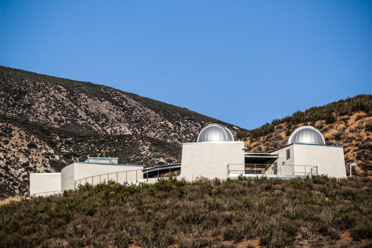 White concrete observatory building with two telescope domes perched atop a chaparral-covered mountain ridge, under a clear blue sky.