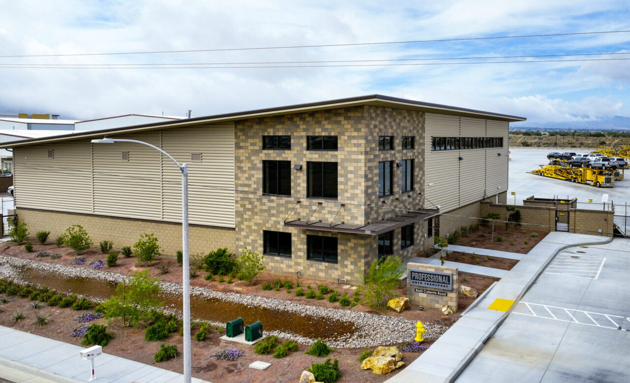 Modern two-story commercial building with beige and brown concrete block walls, metal siding, and large windows, featuring a "Professional Auto Transport" sign at 8660 Caliente Road, with desert landscaping, boulders, and a paved parking lot in the foreground under a partly cloudy sky.