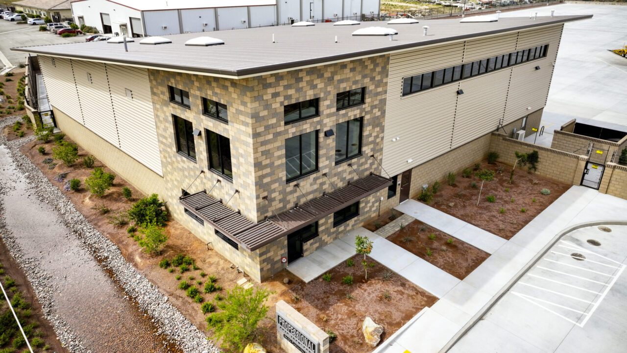 Modern two-story commercial building with beige and brown concrete block walls, metal siding, and large windows, featuring a "Professional Auto Transport" sign at 8660 Caliente Road, with desert landscaping, boulders, and a paved parking lot in the foreground under a partly cloudy sky.