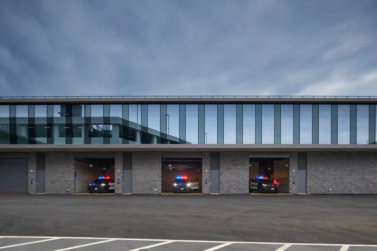 Modern police station building at dusk with a glass-paneled upper floor and gray brick ground level, three open garage bays showing patrol vehicles with lights activated, and an empty parking lot in the foreground.