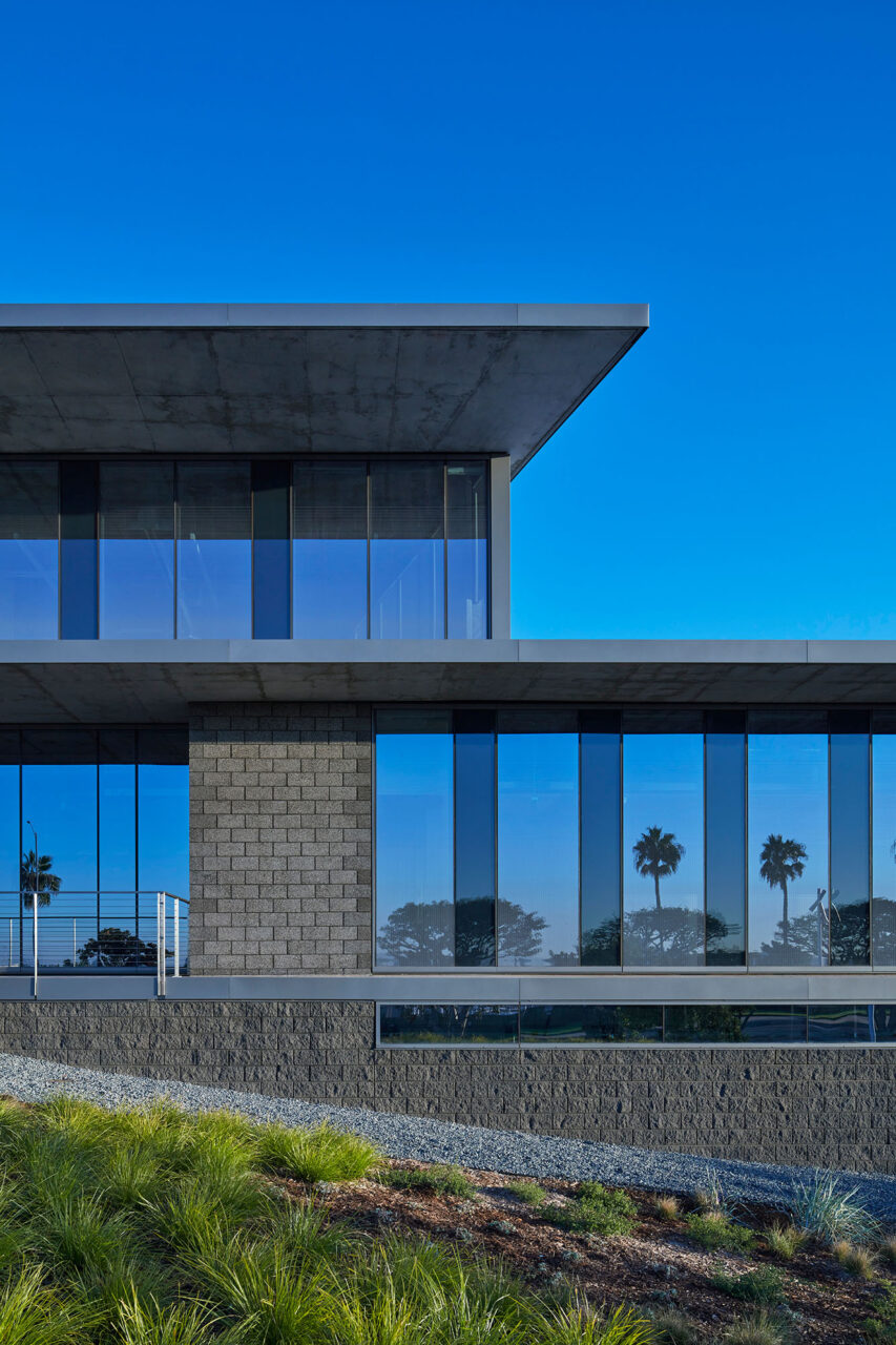 Modern two-story building with cantilevered concrete roof overhangs, floor-to-ceiling glass windows reflecting palm trees, and textured gray block cladding, set above a gravel and native grass landscape slope.
