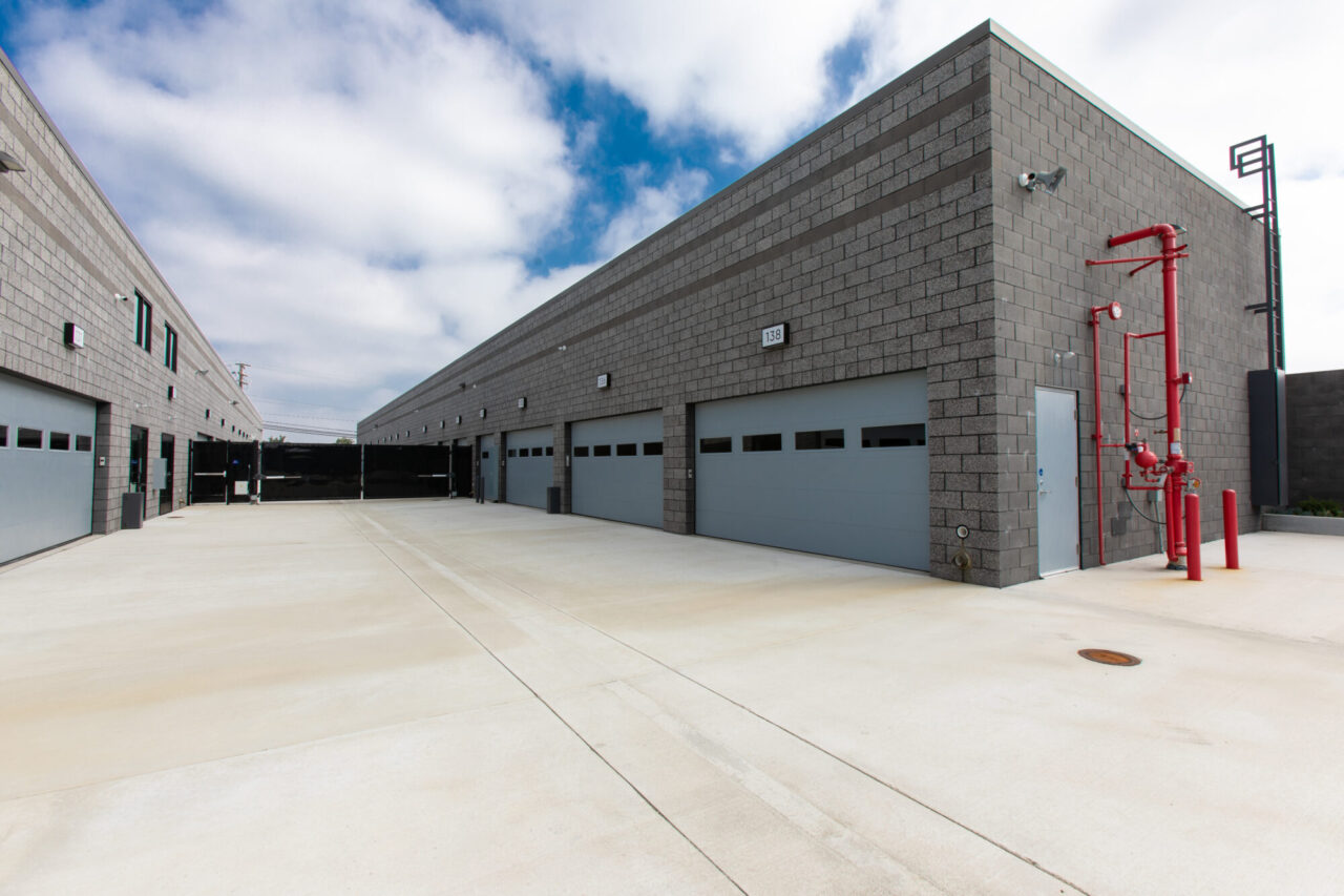 Industrial warehouse building numbered 138 with gray concrete block walls, multiple gray roll-up loading doors, red exterior fire suppression piping, and a wide concrete driveway apron.