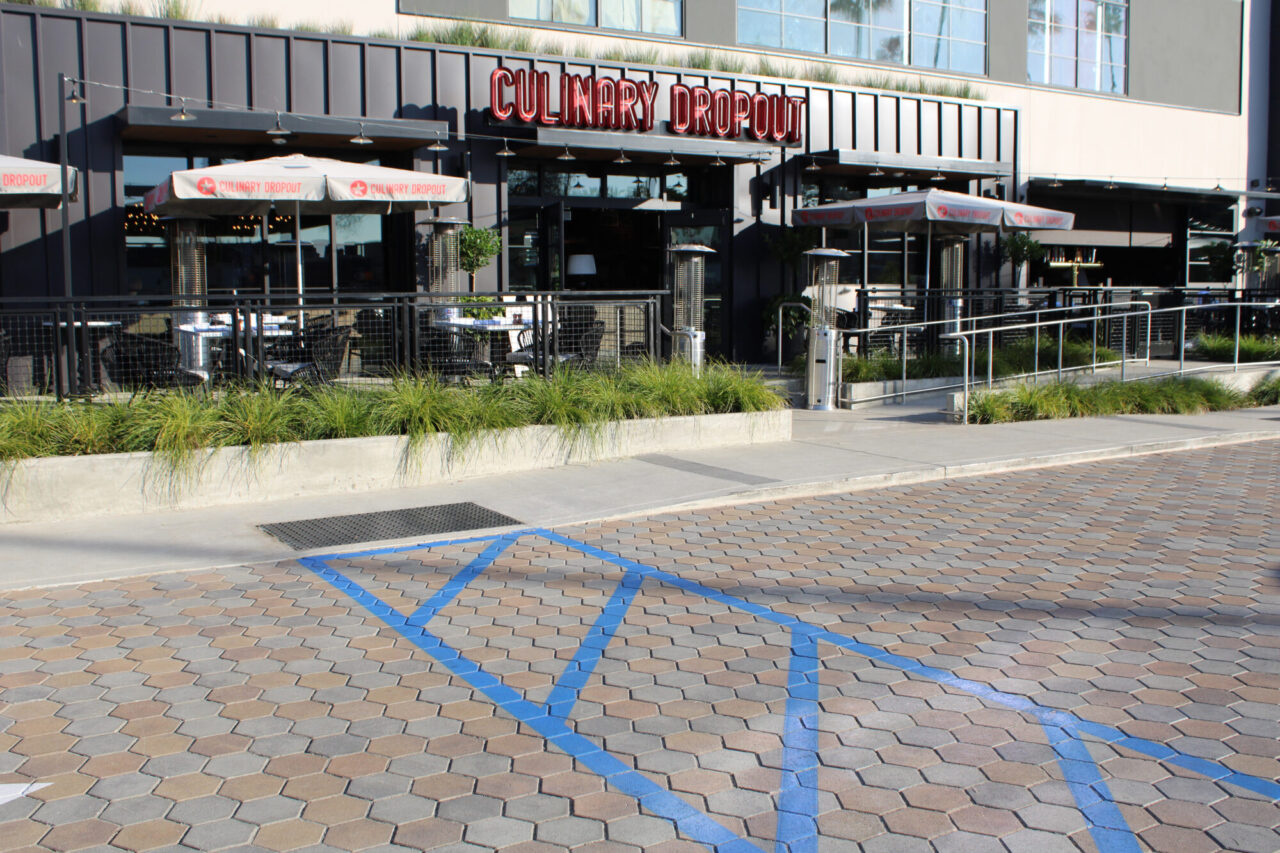 Exterior of the Culinary Dropout restaurant with a red neon sign above a black storefront, outdoor patio seating with umbrellas and heat lamps, grass planters, and a hexagonal paver driveway with blue handicap crossing lines in the foreground.