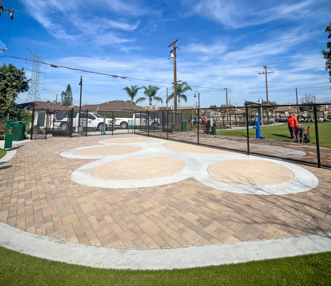 Community dog park plaza with a decorative concrete and paver paw-print design bordered by artificial turf, fenced sports courts and workers in the background, palm trees, and utility poles under a bright blue sky.