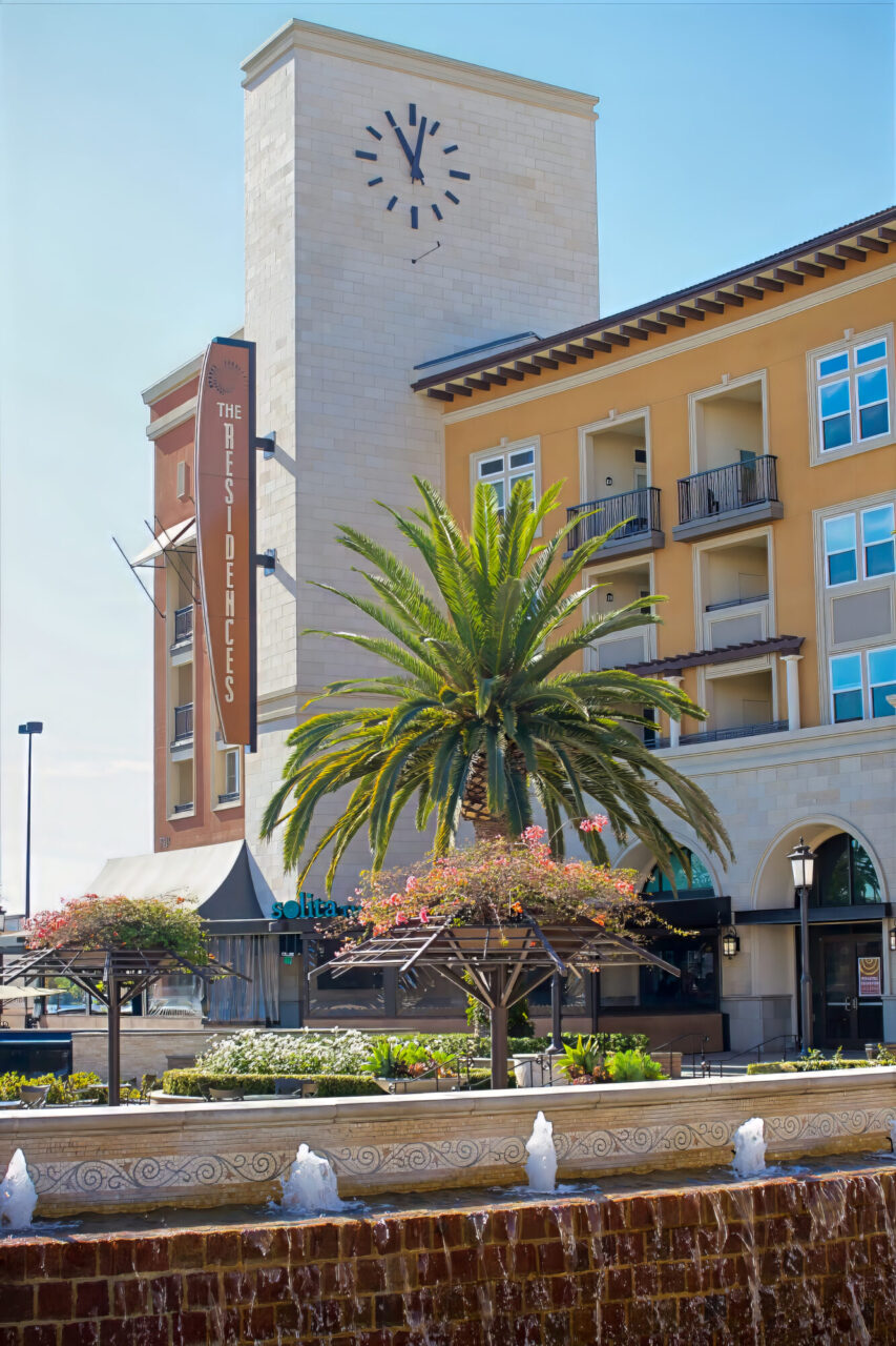 and "The Residences" building sign, a mature palm tree, wood pergolas over an outdoor restaurant patio, and a decorative stone fountain with jets in the foreground.