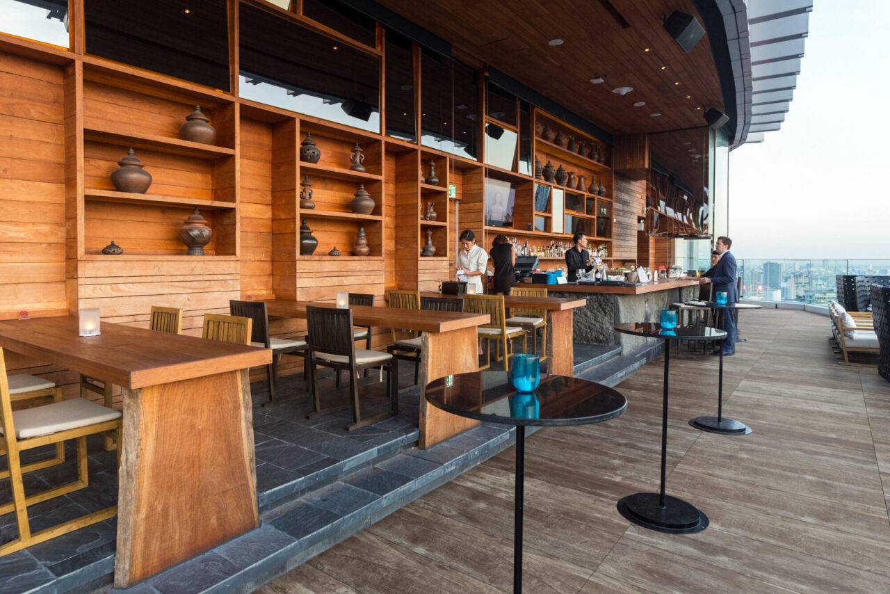 Rooftop bar and lounge with wooden tables, chairs, and shelving displaying decorative pottery, a staffed bar with bartenders and a guest, small round cocktail tables with blue glass candle holders, porcelain plank flooring, and a hazy city skyline visible beyond the open-air railing.