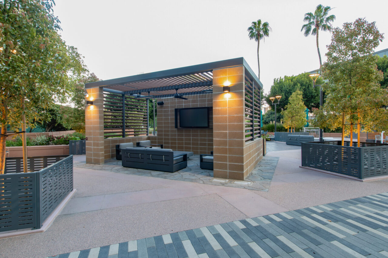 Outdoor lounge cabana with concrete pillars, slatted wood pergola roof, wall-mounted TV, and dark modern sofas, illuminated by sconce lighting, set on a striped paver plaza with planter boxes and palm trees at day.
