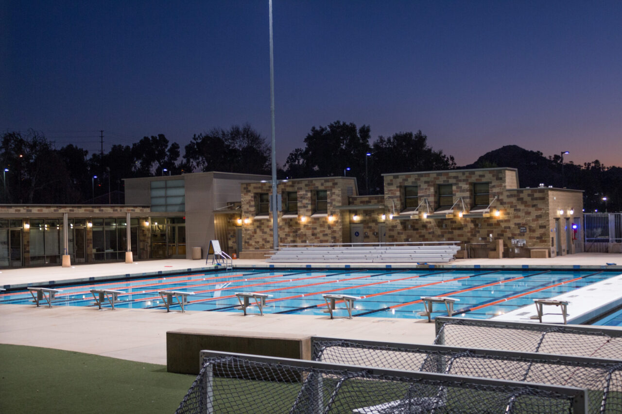 Community aquatic center at dusk with an illuminated Olympic-style lap pool featuring starting blocks and lane lines, bleacher seating, a multicolored concrete block building with lit sconces, and a hillside silhouette in the background.