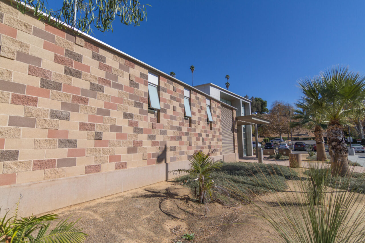 Modern building exterior with multicolored brick facade in tan, rust, and gray tones, angled awning windows, a covered entryway, and drought-tolerant landscaping with palms and desert grasses, with a parking lot visible beyond.
