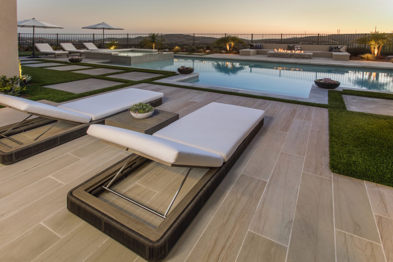 Lounge chairs on a natural stone pool deck with a linear fire feature overlooking hills at dusk.