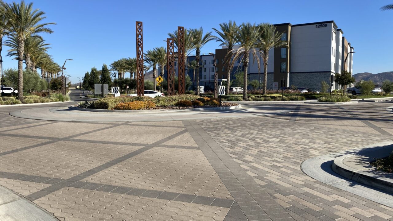 Landscaped roundabout with decorative rusted metal trellis pillars, palm trees, and flowering shrubs, surrounded by a patterned paver street, with a multi-story hotel building numbered 12509 and mountains in the background under a clear blue sky.